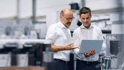 Photograph of two men in technical warehouse in white shirts talking about something that can be seen on a laptop