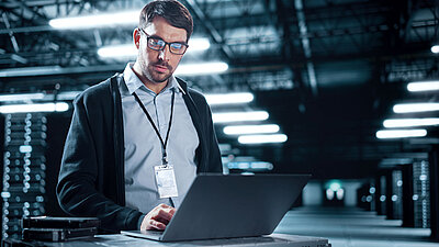 Young IT employee with glasses, three-day beard, light blue shirt, dark blue cardigan and security badge around his neck stands in a huge gray-blue server room and checks security settings on his laptop