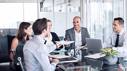 Five people sit at a meeting table and exchange documents