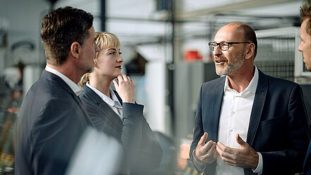 Photograph of a group of business people talking in a production hall: Man on the right with half bald head, glasses and gray beard in jacket and white shirt is explaining something to a younger colleague on the left and an attentively listening blonde colleague