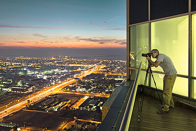 LANCOM SD-WAN: Trustworthy SD-WAN from Germany Photo of a man taking a picture of the illuminated city at night with his camera tripod from the balcony / roof of his penthouse apartment above a big city