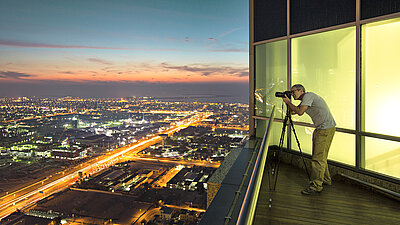 LANCOM SD-WAN: Trustworthy SD-WAN from Germany Photo of a man taking a picture of the illuminated city at night with his camera tripod from the balcony / roof of his penthouse apartment above a big city