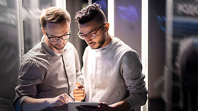 Integrated security. Maximum control. Two men stand in a server room and analyze data together on a tablet.