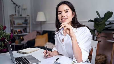 Young brunette woman in a white business blouse smiles contentedly at the camera and holds her recently removed glasses in her hand as she sits at her desk in her home office and takes notes next to her laptop; plants, a floor lamp and a bookshelf can be seen in the background