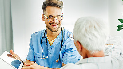 Photograph of a cheerfully laughing, blond, young geriatric male nurse with glasses in a blue doctor's coat and with a stethoscope around his neck, showing only a file to an elderly lady with short white hair, who can be seen in the foreground from behind