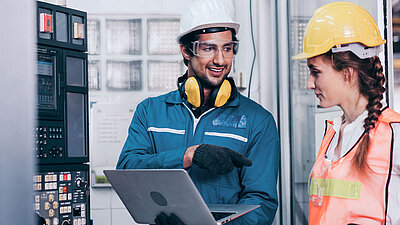 Black-haired worker in utility plant with white hard hat, safety goggles and yellow noise protection headphones in blue overalls and work gloves holding laptop and standing to the right of a control box (left); he explains something to his brunette colleague to his right, smiling; his colleague is also wearing a yellow hard hat and safety vest and listening attentively