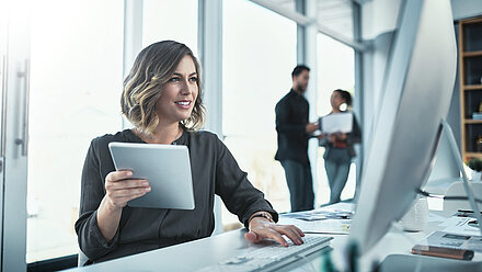 Woman sitting at a desk, holding a tablet and looking at the computer, two colleagues talking in the background