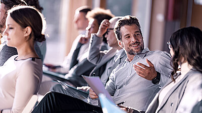 Business people at a seminar or conference sit in a row of chairs. A middle-aged man wearing a gray shirt and a watch on his wrist is smiling and talking to a woman next to him while holding a document in his hand. In the background, other participants in business attire can be seen concentrating on the conversation or taking notes. The image conveys a relaxed and cooperative atmosphere during a business event.