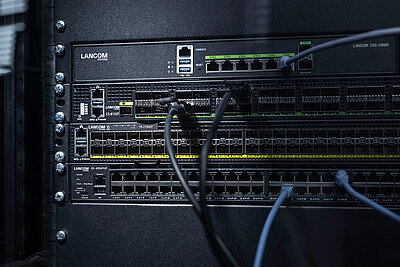 Three LANCOM switches installed in the rack with cables: core switch LANCOM CS-8132F, aggregation switch LANCOM YS-7154CF, and access switch LANCOM XS-4554XUP