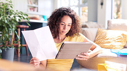 Woman sitting in front of the sofa, holding a tablet in her left hand and a piece of paper in her right hand