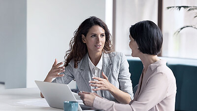 Secure Edge Computing Two colleagues are sitting at a table in front of a laptop, one of them is gesturing with her hands.