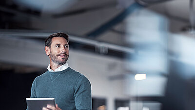 Brunette middle-aged man with a beard and a combination of a blue sweater over a white shirt on the left of the picture stands smiling confidently in modern office space, holding a tablet in his hand and looking inspired and thoughtful up to the right