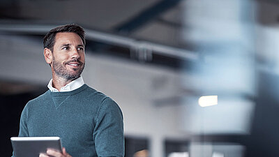 Brunette middle-aged man with a beard and a combination of a blue sweater over a white shirt on the left of the picture stands smiling confidently in modern office space, holding a tablet in his hand and looking inspired and thoughtful up to the right