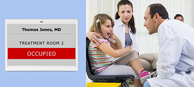 Digital room signage of a treatment room by means of a LANCOM Wireless ePaper Display. A young girl and her mother are sitting in a treatment room facing a doctor. The young girl is pointing at her teeth.